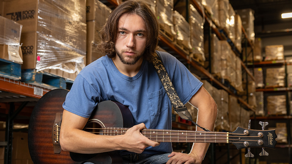 TCC alumnus Christopher Capra holds his bass guitar, flanked by shelves of food the Food Bank of Eastern Oklahoma Warehouse.